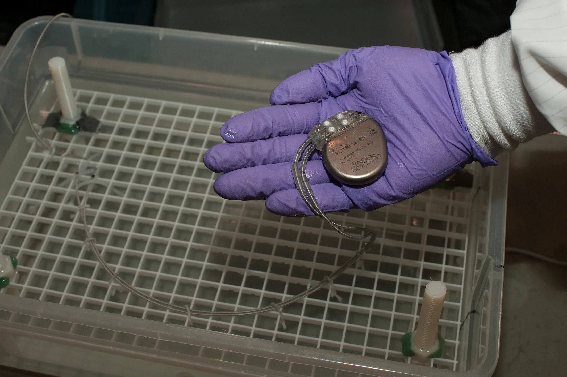 A gloved FDA engineer's hand holding a sealed titanium pacemaker with coiled leads above a laboratory test bath.