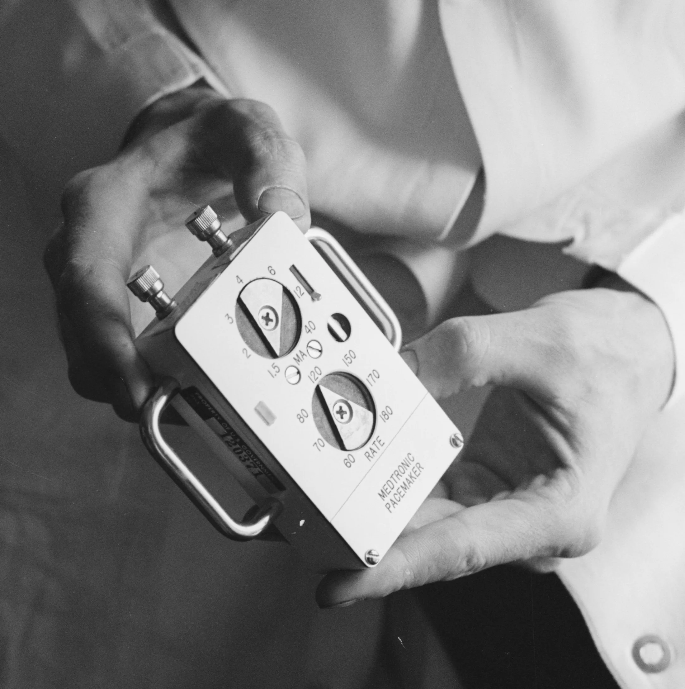 Black-and-white photograph of two hands adjusting the dials of a boxy 1961 Medtronic external pacemaker.